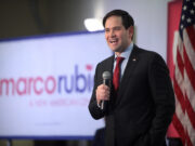 Marco Rubio speaking into a microphone in front of an American flag