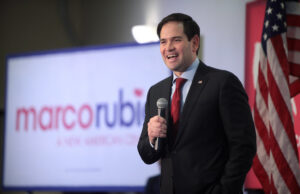 Marco Rubio speaking into a microphone in front of an American flag