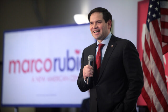 Marco Rubio Marco Rubio speaking into a microphone in front of an American flag